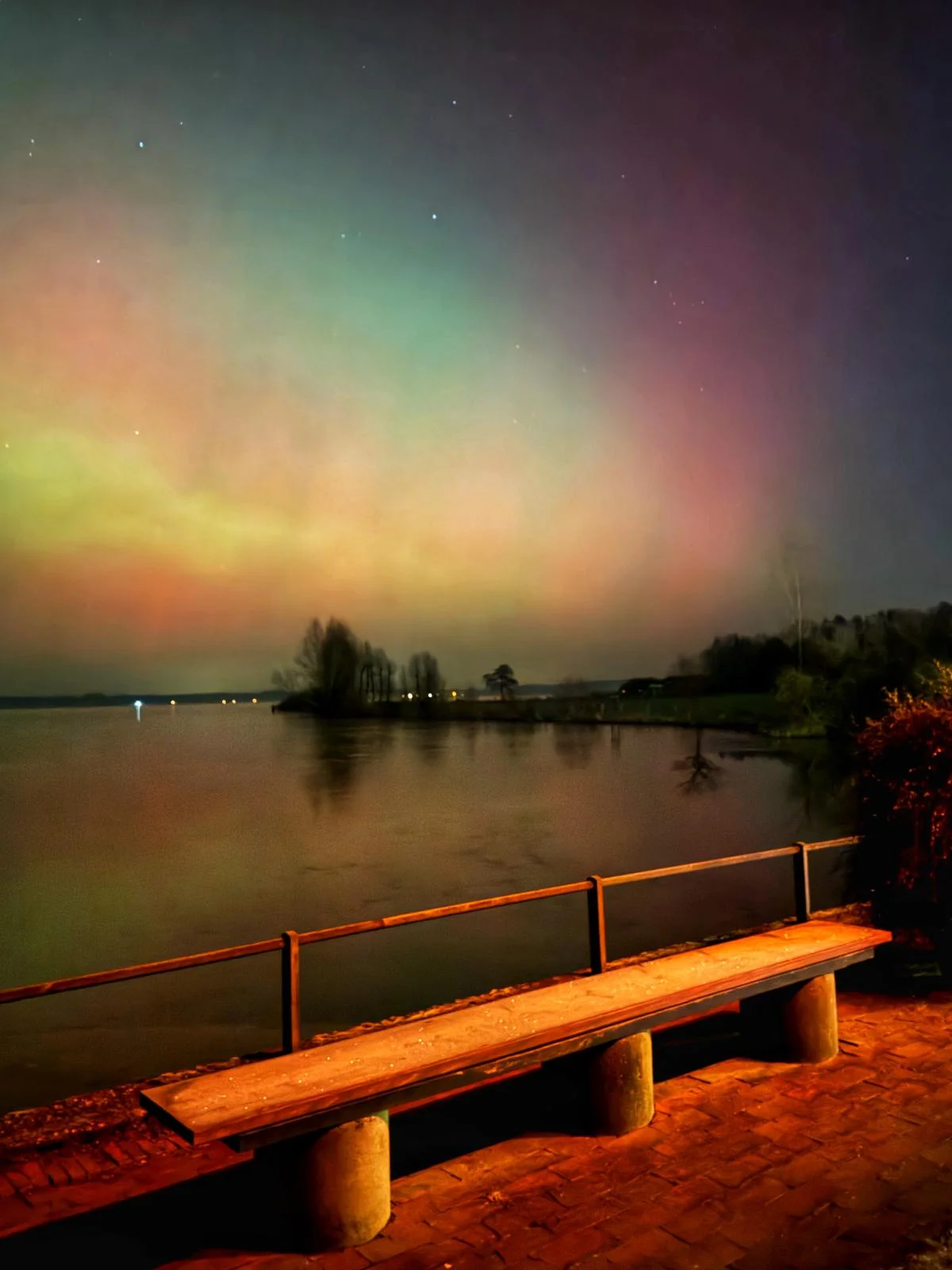 Tanzende grüne und pinke Polarlichter am Nachthimmel über einer Holzbank am See