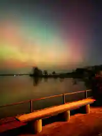 Tanzende grüne und pinke Polarlichter am Nachthimmel über einer Holzbank am See